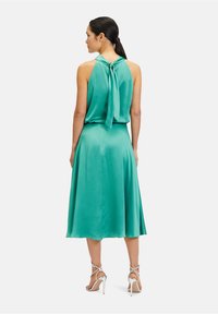 Sukienka z satynowego materiału w kolorze teal z dekoltem halter i kokardą z tyłu, z luźną spódnicą, zestawiona ze srebrnymi sandałkami na obcasie.
