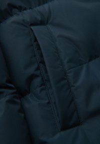 Donker teal pufferjack met een gequiltde textuur, voorzien van een klein ritsvakje. Het materiaal ziet er soepel en waterafstotend uit.