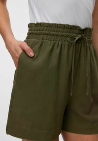 Shorts de color verde oliva, con cintura elástica y cordón, que cuentan con bolsillos laterales y una textura de algodón suave, terminando justo por encima de la rodilla.
