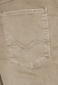 Tejido de mezclilla beige con un bolsillo trasero que presenta un patrón de puntada en zigzag y un pequeño logo plateado cerca de la esquina superior.