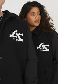 Zwarte jas van een wolmix, met knoopsluiting aan de voorkant en een opvallend wit geborduurd logo op de borst. Gelaagd stof met zwarte accenten.