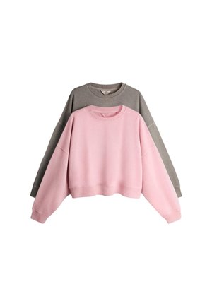 Junge Frau steht vor einer weißen Wand und trägt einen braunen übergroßen Sweatshirt, passende Shorts, weiße Socken und hellblaue Sneaker.