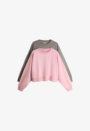 Ung kvinde står mod en hvid væg iført en brun oversized sweatshirt, matchende shorts, hvide sokker og lyseblå sneakers.