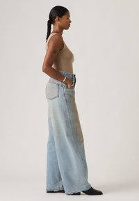 Γυναίκα με πλεγμένα μαλλιά, φορώντας αμάνικο μπεζ τοπ, φαρδιά φαρδιά ανοιχτό μπλε τζιν παντελόνια, μαύρα παπούτσια και ασημένια βραχιόλια, ποζάρει σε προφίλ.