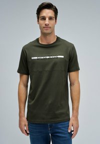 Joven de pie vistiendo una camiseta verde oliva con el texto "Salsa Jeans" y pantalones vaqueros azules frente a un fondo gris claro liso.