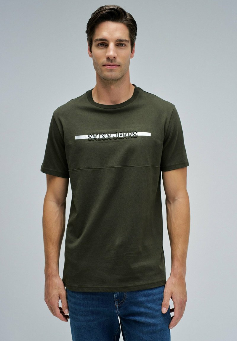 Joven de pie vistiendo una camiseta verde oliva con el texto "Salsa Jeans" y pantalones vaqueros azules frente a un fondo gris claro liso.