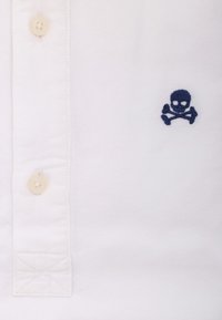 Weißes Hemd mit Knopfverschluss und einem kleinen, navyblauen gestickten Totenkopf- und Kreuzbones-Design. Das Material wirkt glatt mit einer weichen Textur.