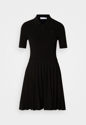Black ribbed polo dress with short sleeves, featuring a zip neckline and pleated skirt. Made from soft fabric, designed for a fitted silhouette.