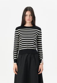 Jeune femme avec de longs cheveux bruns portant un haut à rayures noires et blanches à manches longues et une jupe noire, se tenant devant un fond blanc.