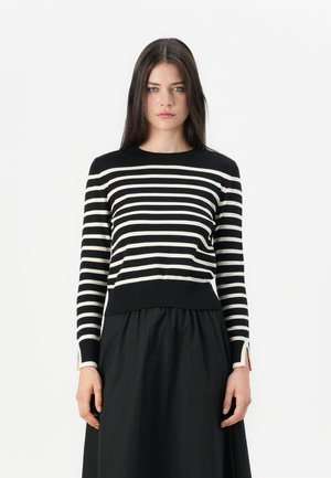 Jonge vrouw met lang zwart haar, gekleed in een zwart-wit gestreepte lange mouwen top en een zwarte rok, staande tegen een witte achtergrond.