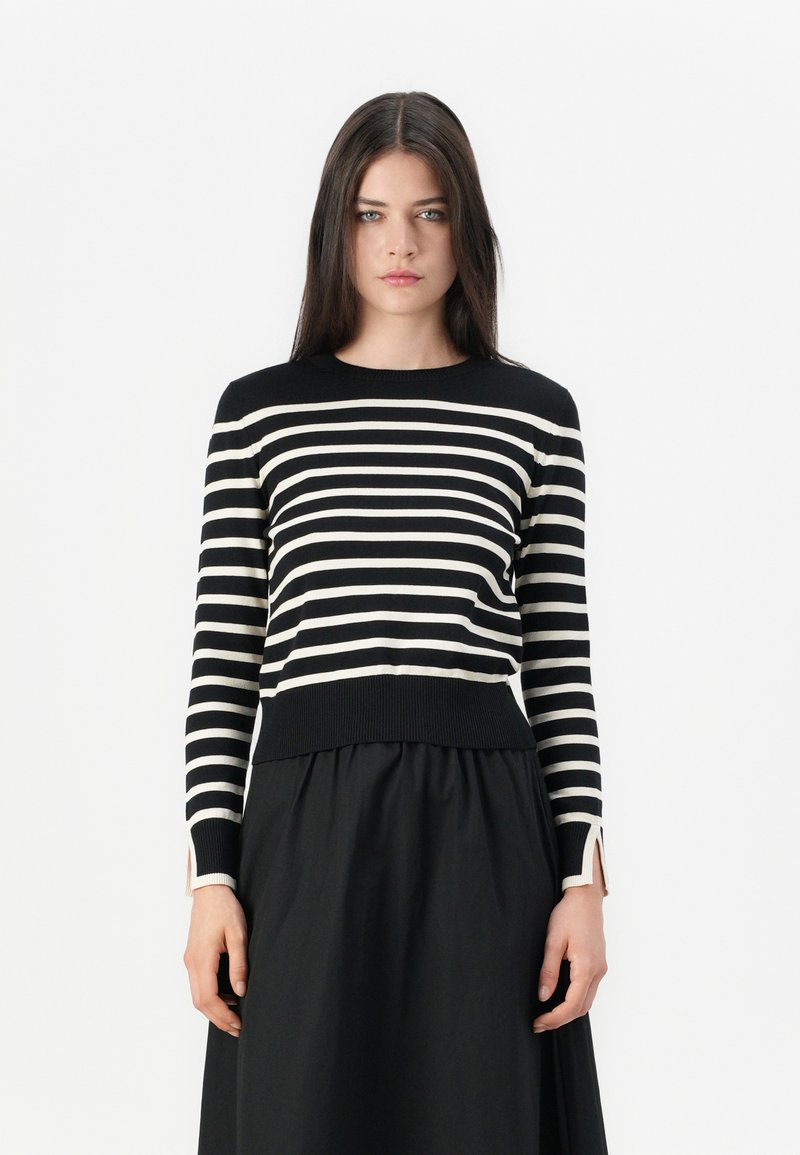 Jeune femme avec de longs cheveux bruns portant un haut à rayures noires et blanches à manches longues et une jupe noire, se tenant devant un fond blanc.