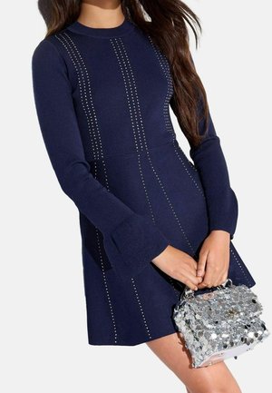 Donna che indossa un abito blu navy a maniche lunghe con dettagli verticali di borchie, mentre tiene davanti a sé una piccola borsa argentata di paillettes.