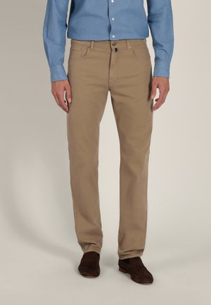 Mann trägt beige gerade geschnittene Hose, blaues Hemd mit Knöpfen und dunkelbraune Slipper, steht vor einfachem Hintergrund.