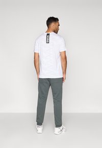 T-shirt de camuflagem branca com um rótulo preto "BORG" nas costas, combinada com calças de fato cinzas e ténis brancos.