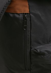 Fermeture éclair en tissu noir, avec un tirette sombre et une bandoulière marron contrastante. Surface texturée avec un détail de poche latérale.