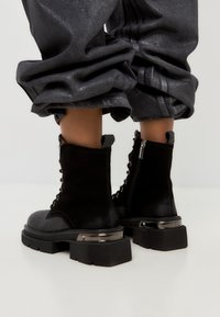 Schwarze Schnürstiefeletten mit einer strukturierten Oberseite, robuster schwarzer Sohle, metallischen Akzenten an der Ferse und einem seitlichen Reißverschluss.