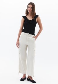 Musta hihaton toppi, jossa on röyhelöyksityiskohta, yhdistettynä kermaisiin cargo-housuihin, joissa on sivutaskut ja nauhat hemissä, käytetty mustien kenkien kanssa.