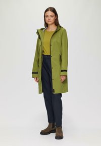 Zielony wodoodporny płaszcz przeciwdeszczowy z kapturem, czarnymi akcentami i zapięciami na zatrzask, noszony na musztardowej bluzce i ciemnych spodniach, w połączeniu z brązowymi botkami za kostkę.