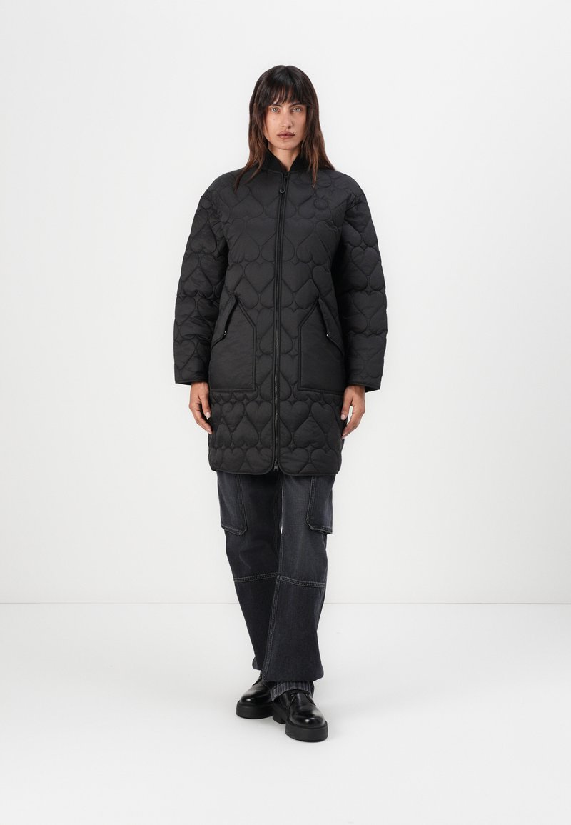 Zwart gewatteerd lang jasje met een rits aan de voorkant, hoge kraag, twee zijzakken en een ontspannen pasvorm, gecombineerd met donkere baggy broeken en chunky schoenen.