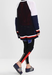 Meshstickad tröja med marinblå, vit och röd rand; ihopparad med svarta leggings med röda och blå detaljer samt siddragkedjor; rutiga slidesandaler.