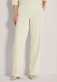 Vrouw in crèmekleurige geribbelde wijde broek met handen in de zakken en beige puntige hakken tegen een lichtgrijze achtergrond.