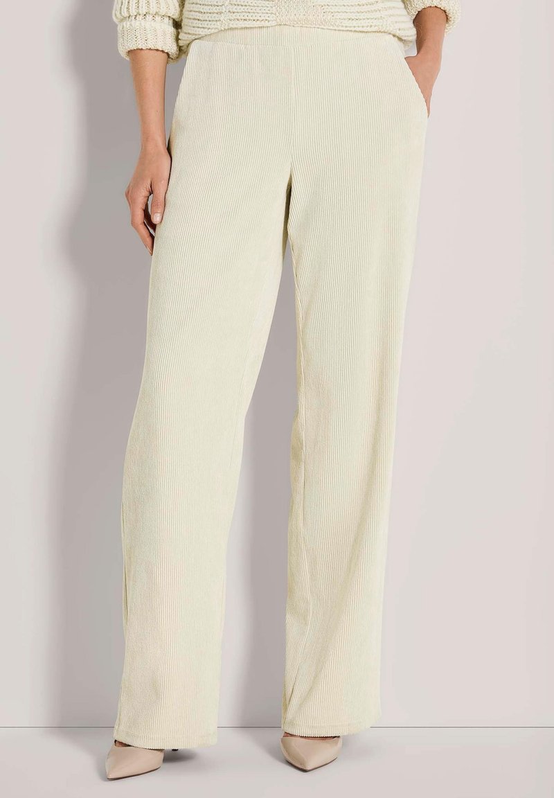 Vrouw in crèmekleurige geribbelde wijde broek met handen in de zakken en beige puntige hakken tegen een lichtgrijze achtergrond.