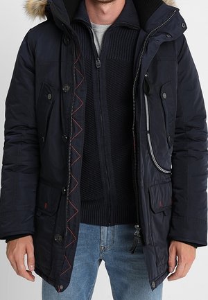 Homme portant un parka d'hiver bleu marine avec capuche bordée de fourrure, un pull noir à fermeture éclair en tricot, et un jean bleu clair, debout avec les mains détendues.