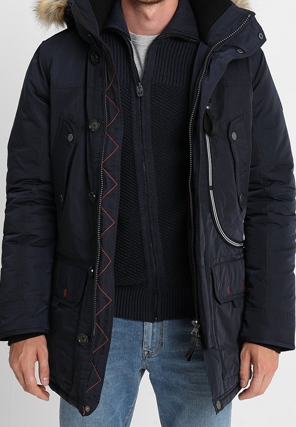 Homme portant un parka d'hiver bleu marine avec capuche bordée de fourrure, un pull noir à fermeture éclair en tricot, et un jean bleu clair, debout avec les mains détendues.