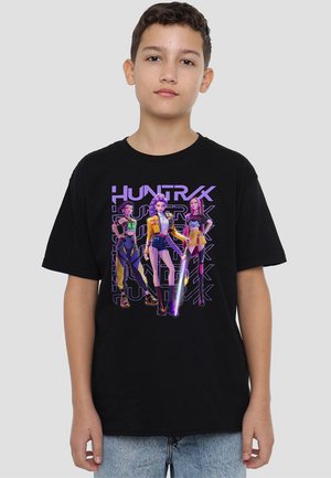 Junge in schwarzem T-Shirt mit drei animierten weiblichen Figuren und dem Text "HUNTRX" in Lila auf einem hellgrauen Hintergrund.