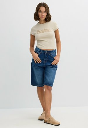 Camiseta ajustada beige con texto marrón, combinada con pantalones cortos de mezclilla azul hasta la rodilla y zapatos de slip-on beige con un acento texturizado.