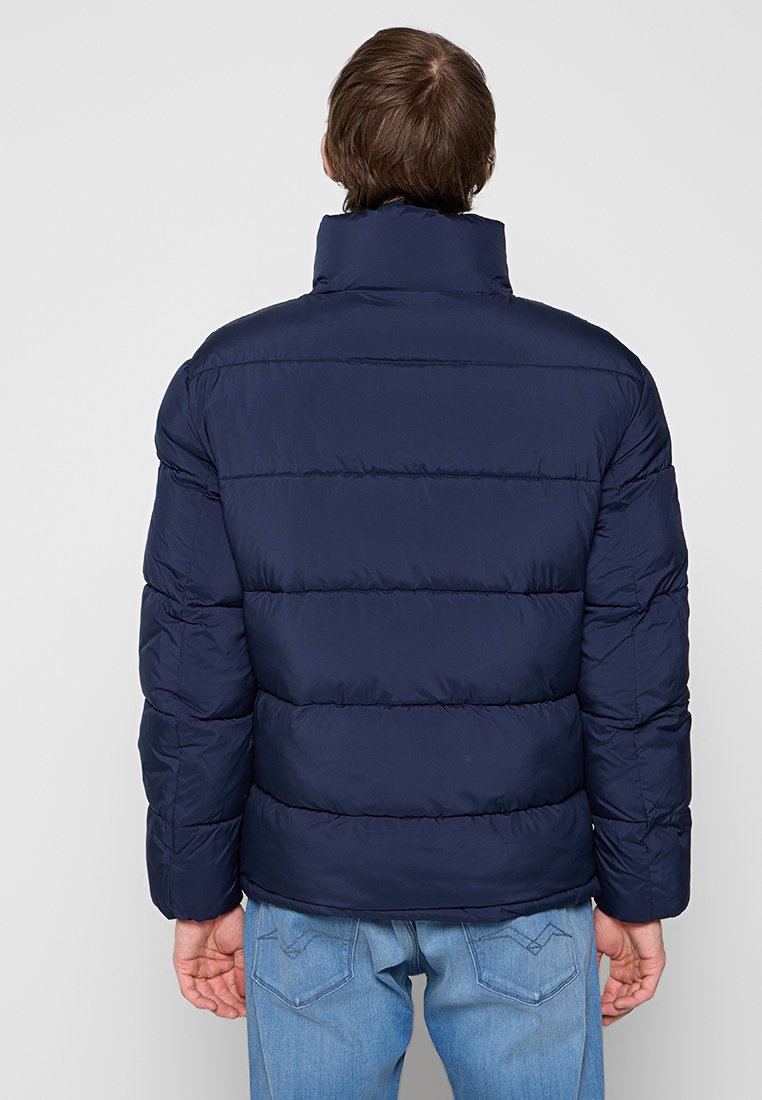 Mężczyzna w granatowej kurtce puchowej i jasnoniebieskich dżinsach, stojący tyłem na jednolitym jasnoszarym tle.