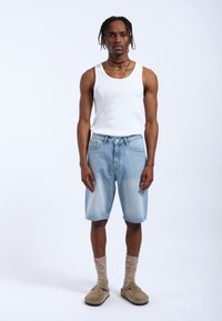 Lichtblauwe denim shorts met een relaxed fit, gecombineerd met een witte tanktop, beige instapschoenen en gestreepte bruine sokken.
