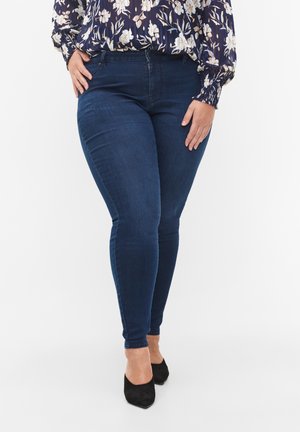 Femme portant un jean skinny bleu foncé, des talons noirs et un chemisier bleu marine à manches longues avec un motif floral blanc, debout les mains près des poches.