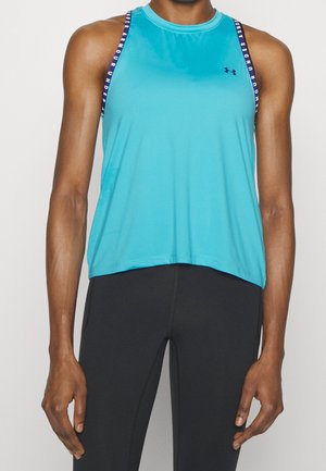 Athletische Person, die ein ärmelloses türkisfarbenes Under Armour Top und schwarze Leggings trägt, steht mit locker an den Seiten hängenden Armen.