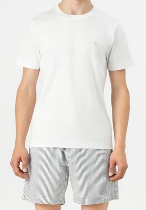 Mand iført en almindelig hvid kortærmet t-shirt og grå og hvide lodrette stribede shorts, stående med armene afslappet ned langs siderne.