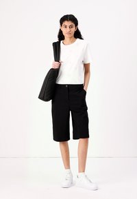 T-shirt en coton blanc avec un motif à œillets, shorts noirs jusqu'aux genoux et baskets blanches. Le mannequin tient un sac à bandoulière matelassé noir.