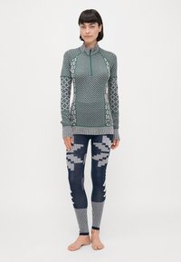 Langermet topp i grønn med mønstrete design og høy krage, kombinert med marineblå leggings som har geometriske detaljer og stripete elementer.
