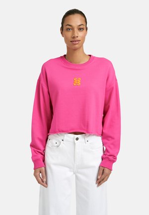 Woman wearing a bright pink cropped sweatshirt with an orange logo on the chest and high-waisted white jeans against a plain background.