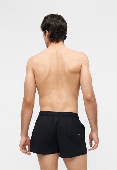 Άντρας με κοντά σκούρα μαλλιά στέκεται γυρισμένος, χωρίς πουκάμισο, φοράει μαύρο μαγιό μπροστά σε απλό φωτεινό φόντο.