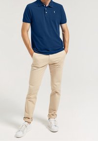 Námořnická modrá pólová košile s límečkem, obsahující tři knoflíky a malé logo. V kombinaci s béžovými kalhotami a bílými teniskami.