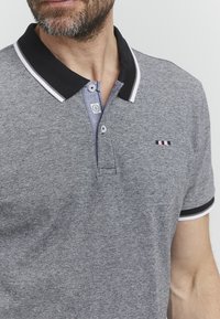 Sivi polo majica iz teksturiranega blaga, ki ima črn ovratnik in bele detajle, zapenjanje na dva gumbka ter majhen logo na prsih.