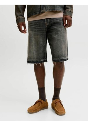 Mann trägt ausgewaschene schwarze Jeans-Shorts mit ausgefransten Säumen, braune Wildlederschuhe, dunkle Socken, helles Hemd und Jeansjacke vor weißem Hintergrund.