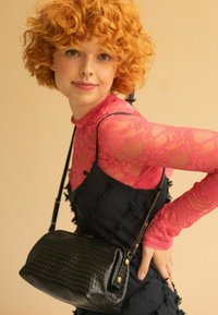 Borsa a tracolla intrecciata nera con dettagli dorati. Modella con una maglietta a maniche lunghe in pizzo rosa indossata sopra un vestito nero testurizzato.