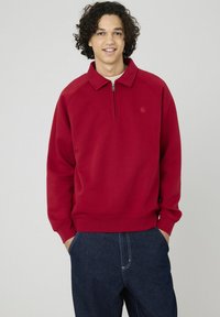 Sweat-shirt rouge avec un col zippé, des poignets et un ourlet côtelés. Le tissu semble lisse avec une coupe décontractée. Porté sur un pantalon en denim bleu.