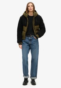 Zwarte en olijfgroene pufferjack met capuchon, gedragen over een zwarte kabelgebreide trui. Een paar opgerolde blauwe jeans en zwarte laarzen.