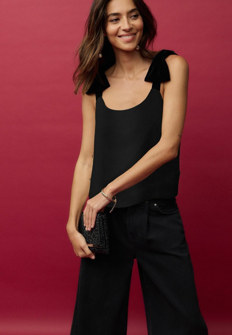 Top negru fără mâneci cu accente de catifea pe umeri, asociat cu pantaloni negri largi. Modelul ține o poșetă neagră cu mărgeluțe. Fundalul este de culoare roșie uniformă.