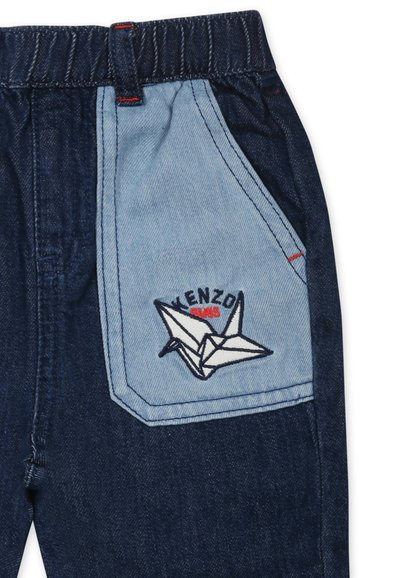 Pantalon en denim avec une pièce lumineuse bleue, des coutures rouges et une grue en origami brodée en blanc avec "KENZO" en noir et rouge au-dessus.
