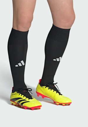 Des crampons de football jaunes vifs avec des accents noirs, présentant un design texturé et une semelle rouge ; associés à des chaussettes noires jusqu'aux genoux affichant un logo.