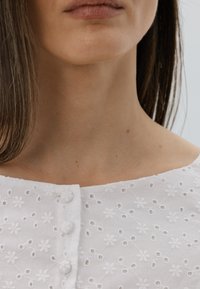 Biała bluzka haftowana z kwiatowymi wzorami, z zapięciem na guziki z przodu oraz szerokim, kwadratowym dekoltem. Materiał jest lekki i teksturowany.