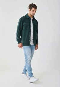 Camicia in flanella verde scuro con chiusura a bottoni e due tasche sul petto, indossata sopra una maglietta bianca, abbinata a jeans azzurri chiari e sneakers bianche.